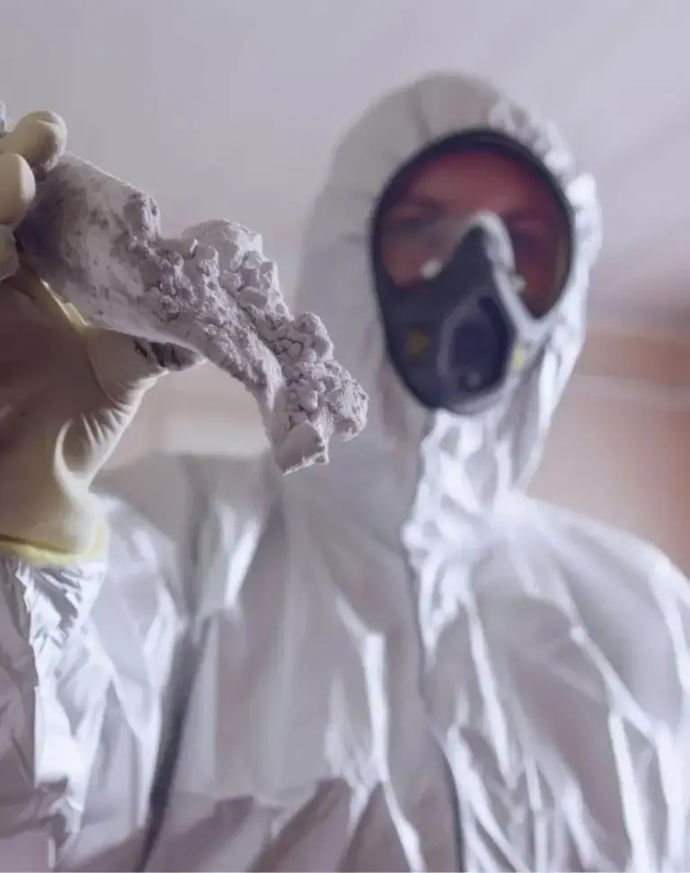 A gloved hand holding a broken piece of grey asbestos lagging up to the camera, with a masked operative out of focus behind it.