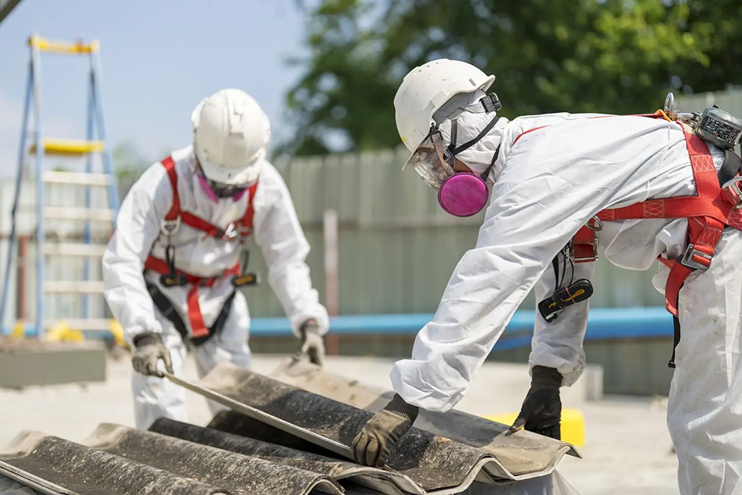Two operatives in coveralls, respirators and fall-arrest harnesses lowering a whole corrugated cement sheet by hand rather than breaking it.